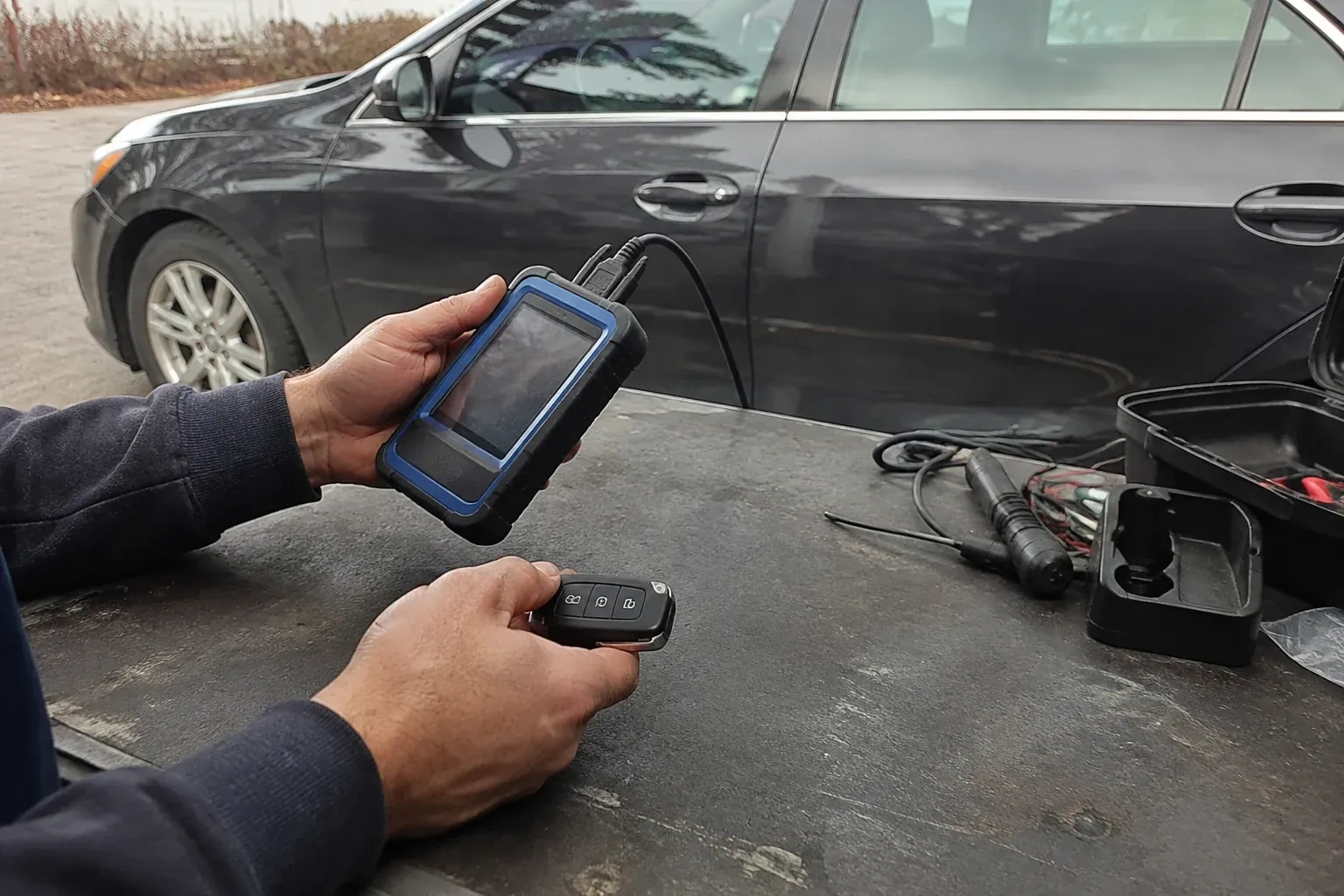 Hands holding a blue-edged electronic device and car-key fob at a worktable beside a parked car. — Transponder Key Programming, Richmond Hill