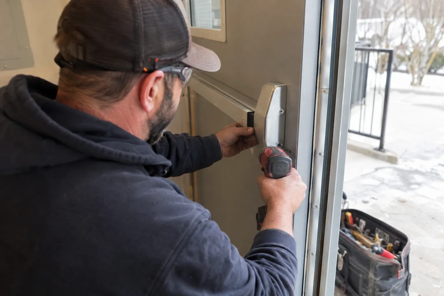 Technician in a cap and safety glasses holding a push bar end cap against a metal door with a red drill. — Elgin Mills