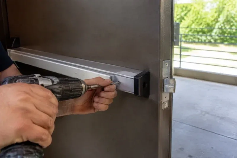 Hands using a power driver at the end of a horizontal push bar on an open metal door. — Panic & Exit Hardware, Richmond Hill